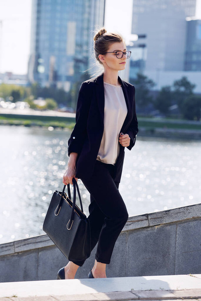 Young elegant businesswoman with black leather handbag moving upstairs in front of camera against riverside and group of modern buildings - Foto, Imagen