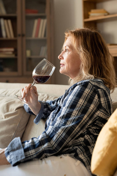 serene volwassen vrouw in casualwear met een glas rode wijn terwijl zitten op de bank in huis omgeving en genieten van rust in de avond - Foto, afbeelding