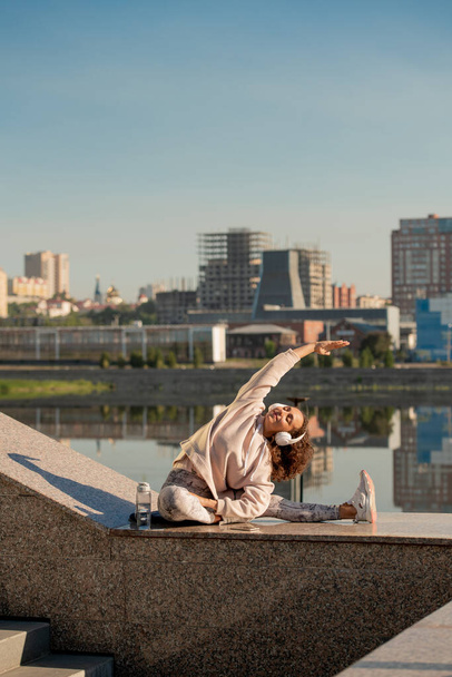Young brunette sportswoman in activewear and headphones stretching leg while exercising on marble structure in urban environment - Photo, Image