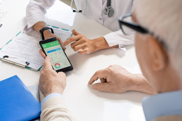Hands of young female dietitian or nutritionist holding smartphone over medical document and showing her senior patient important data - Foto, Imagen