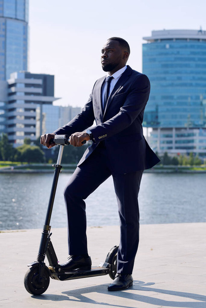 Young elegant businessman in formalwear standing on electric scooter by riverside against group of tall modern buildings on sunny day - Foto, Bild
