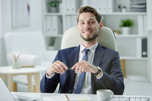 Uśmiechnięty przystojny biznesmen ze ścierniskiem siedzący przy biurku i słuchający partnera biznesowego podczas spotkania z nim w biurze - Zdjęcie, obraz