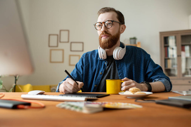 Mladý kreativní mužský návrhář na volné noze, který se napije, zatímco drží stylus nad tabletem a kreslí náčrty nového loga nebo webové stránky - Fotografie, Obrázek