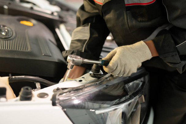 Handschuh-Hand eines jungen Arbeiters des zeitgenössischen Auto-Wartungszentrums in Uniform mit Handgerät, während er Details des Motors während der Reparatur repariert - Foto, Bild