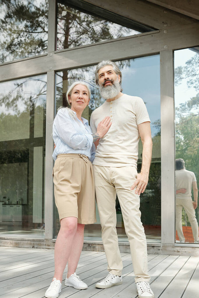 Beautiful senior couple standing against glass cottage and embracing each other outdoors - Photo, Image