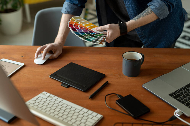 Руки креативного разработчика программного обеспечения или веб-дизайнера с цветовой палитрой, нагибающейся на деревянный стол перед компьютером во время работы - Фото, изображение