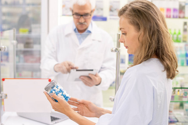 Junge blonde Apothekerin schaut sich Informationen über große Tablettenflaschen an, während sie Informationen über neue Medikamente lernt - Foto, Bild
