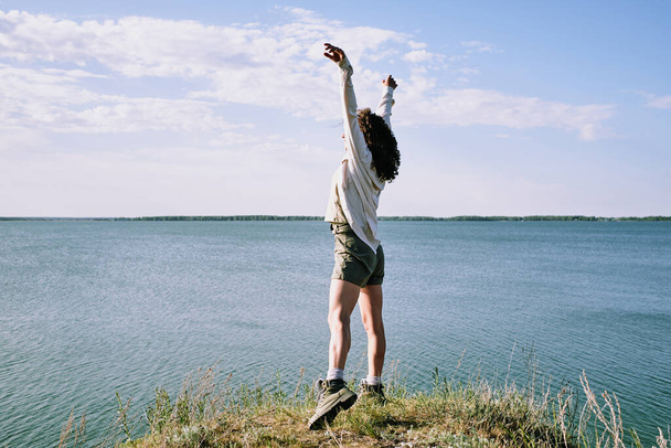 Беззаботная молодая женщина-туристка с вьющимися волосами, стоящая с поднятыми руками и ловящая солнечный свет на берегу - Фото, изображение