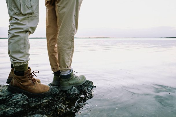 Крупный план неузнаваемой пары в пешеходных сапогах, стоящей на скале и смотрящей на водный пейзаж - Фото, изображение