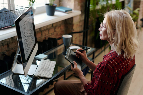Szőke érett női mobil alkalmazás fejlesztő ül asztalnál előtt számítógép monitor és rajz vázlatok táblagépen Stylus - Fotó, kép