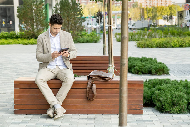 Joven empresario contemporáneo en ropa formal sentado en un banco de madera en el parque y desplazándose en tableta digital en el entorno urbano - Foto, Imagen