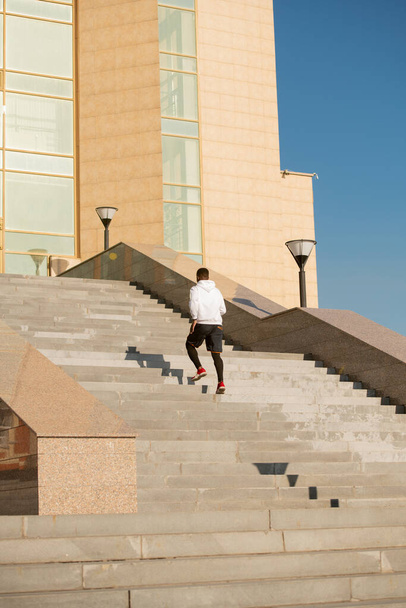 Aktif giyimli genç Afrikalı sporcu şehir ortamında modern mimarinin önündeki büyük merdivenlerin kenarındaki mermer parmaklıklarda oturuyor. - Fotoğraf, Görsel
