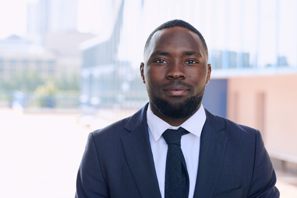 Νεαρός Αφρικανός επιχειρηματίας με κομψό κοστούμι και γραβάτα στέκεται μπροστά στην κάμερα ενάντια σε μια ομάδα σύγχρονων κτιρίων στο αστικό περιβάλλον - Φωτογραφία, εικόνα