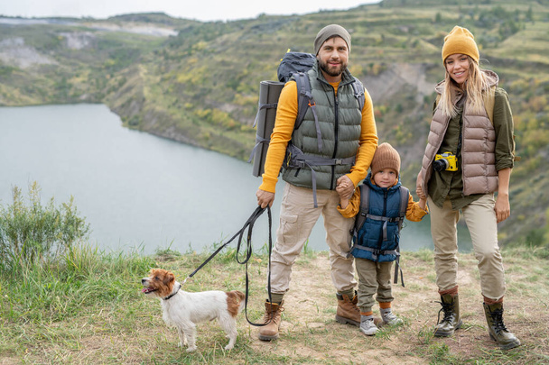 Χαρούμενη νεαρή οικογένεια τριών ατόμων με ζεστά ρούχα και το μικρό σκυλί τους στέκεται στο έδαφος κατά μήκος του ποταμού και των βουνών κατά τη διάρκεια του ταξιδιού - Φωτογραφία, εικόνα