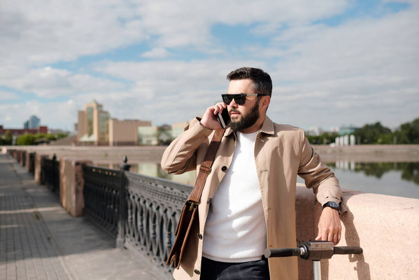 Бородатий бізнесмен в бежевому траншеї, білий в'язаний пуловер і сонцезахисні окуляри розмовляють на смартфоні, стоячи на березі річки
 - Фото, зображення
