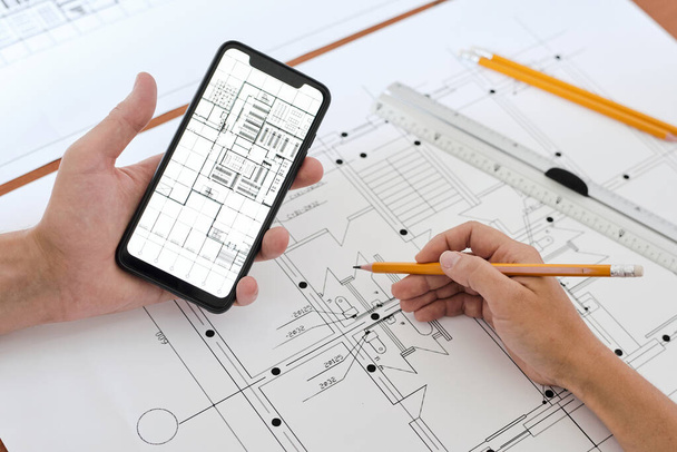Руки молодого занятого инженера или архитектора с карандашом над эскизом нового здания, используя смартфон для исправления или проверки деталей - Фото, изображение