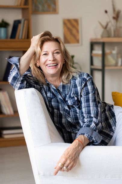 Starší smějící se žena v neformálním oblečení, dívá se na vás se zubatým úsměvem, zatímco sedí v bílém křesle před kamerou v obývacím pokoji - Fotografie, Obrázek