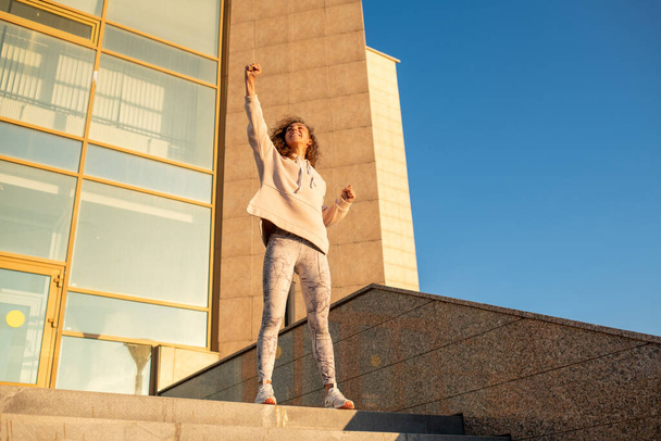 Jonge vrolijke sportvrouw in activewear houdt een been gebogen in de knie terwijl ze op de top van de trap staat tegen de moderne architectuur - Foto, afbeelding