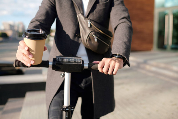 Hände eines eleganten Geschäftsmannes im Mantel halten ein Glas Kaffee in der Hand und fahren auf einem Elektroroller in der städtischen Umgebung, während sie zur Arbeit eilen - Foto, Bild