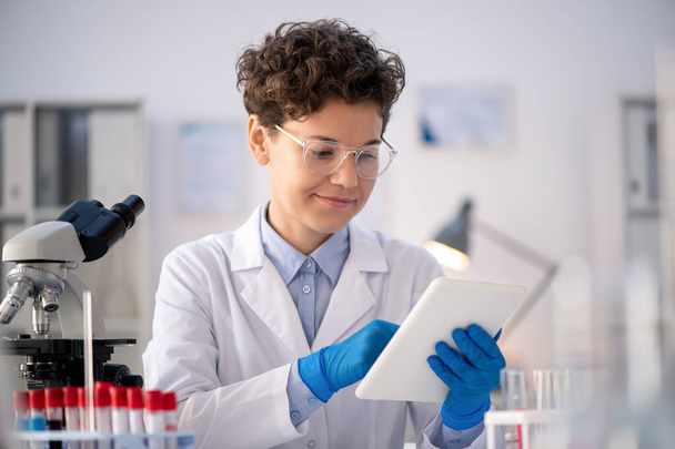 Feliz joven científica contemporánea o farmacéutico en guantes, whitecoat y gafas mirando a través de la información en línea - Foto, Imagen