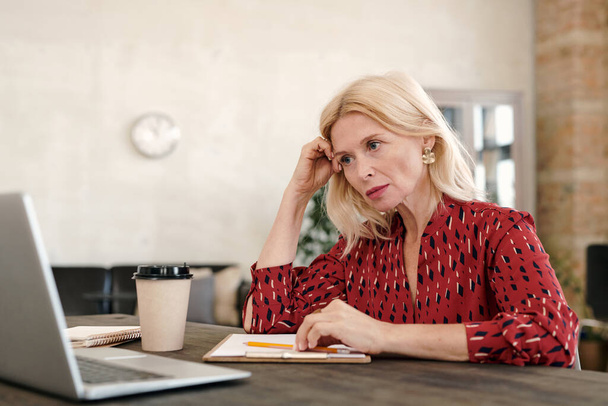 Femme d'affaires blonde mature sérieuse assise devant un ordinateur portable et regardant les données en ligne exposées pendant le réseau dans l'environnement de bureau - Photo, image