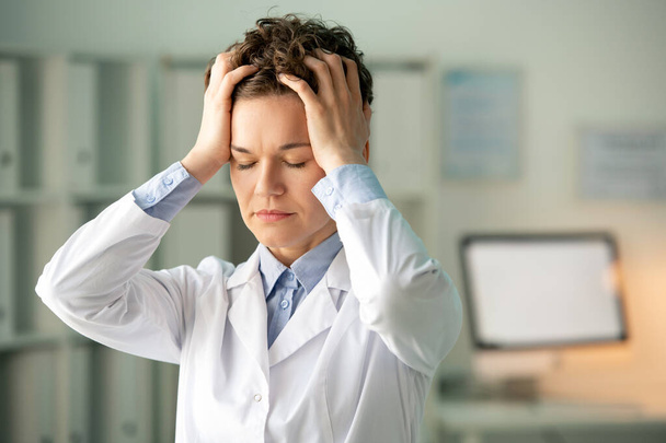 Fiatal fáradt női vegyész vagy gyógyszerész fehérköpenyben megérinti a fejét, miközben a kamera előtt áll a munkahelyen a laboratóriumban - Fotó, kép