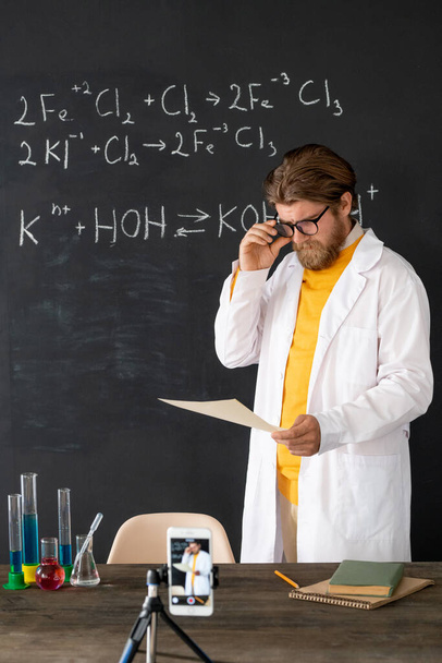 Současný učitel v whitecoat dělá záznam z hodiny chemie pro své online publikum na smartphone kamery proti tabuli - Fotografie, Obrázek