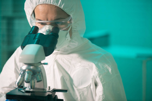 Investigador contemporáneo en batas protectoras, respiradores y anteojos mirando al microscopio mientras estudia nuevas sustancias químicas - Foto, Imagen