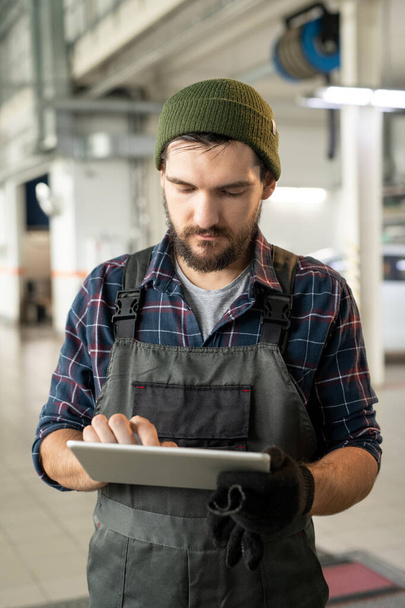Young bearded repairman of contemporary car service touching display of digital tablet while scrolling through online questions of clients - Photo, Image