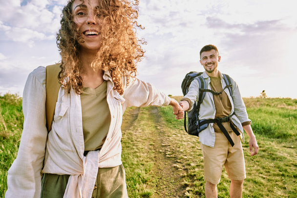 秋に一緒にハイキングをしながら、手を握って田舎道を移動する袋を持つ陽気な若いカップル - 写真・画像