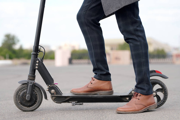 Side view of legs of businessman in brown leather boots and checkered pants standing on electric scooter in urban environment on road - Foto, Imagem