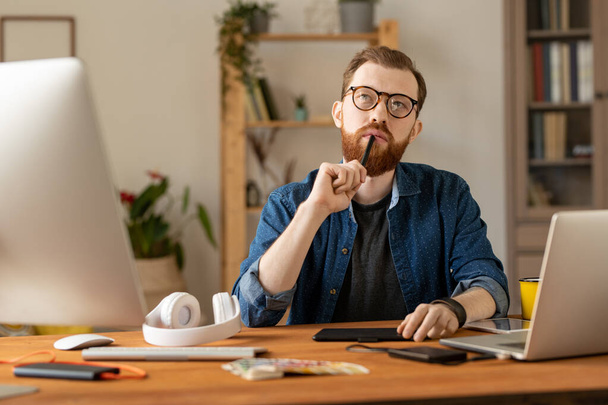 Mladý vousatý zamyšlený freelance designér s stylus u úst sedí u stolu před notebookem, zatímco pracuje přes nové webové stránky doma - Fotografie, Obrázek
