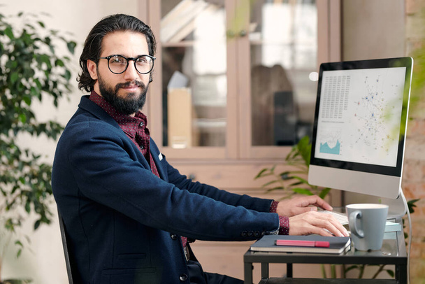 Mladý úspěšný makléř nebo ekonom ve formálním oblečení sedí u stolu před monitorem počítače a dívá se na vás v kancelářském prostředí - Fotografie, Obrázek