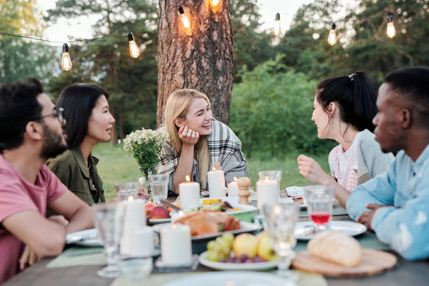 Jóvenes parejas alegres sentadas junto a la mesa festiva bajo el pino, hablando, riendo y disfrutando de sabrosa comida en la cena o fiesta al aire libre - Foto, imagen