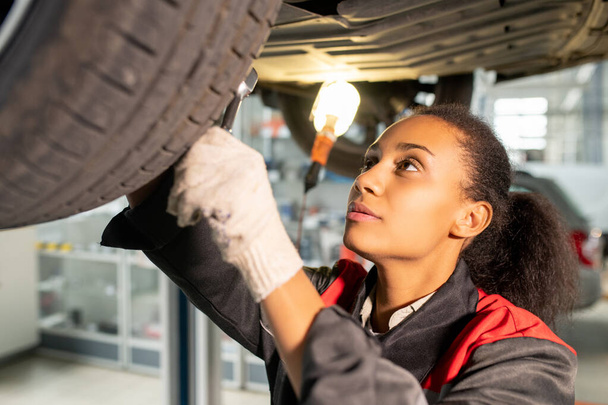 Young female mechanic in gloves and uniform fixing tyre or checking up parts of car bottom while working underneath in workshop environment - Foto, imagen