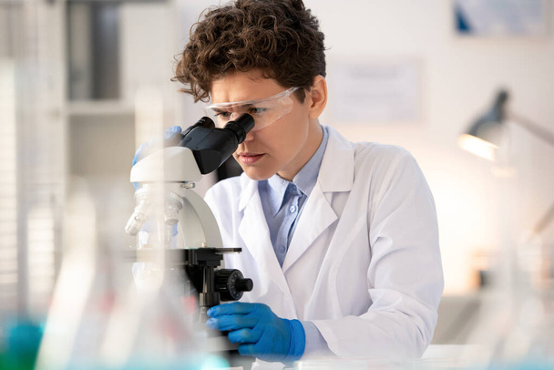 Молодой серьезный ученый или лаборант в перчатках и белых халатах, нагибающийся под микроскопом и изучающий химические вещества - Фото, изображение