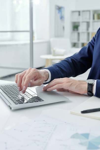 Close-up de empresário irreconhecível em jaqueta sentado na mesa com bloco de notas e digitando no laptop no escritório - Foto, Imagem