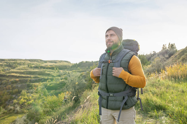 Šťastný mladý muž batohem v teplé casualwear pohybující se po stezce podél hory a zároveň se těší víkendový výlet v přírodním prostředí - Fotografie, Obrázek