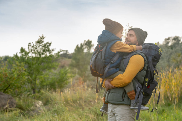 Молодий ніжний батько з рюкзаком тримає свого милого маленького сина на руках проти природного середовища з горою, зеленою травою і кущами
 - Фото, зображення