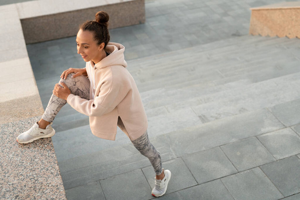 Jovem mulher morena em leggins cinza, cross-shoes e capuz bege mantendo a perna de joelho na telha de mármore durante o treino ao ar livre - Foto, Imagem