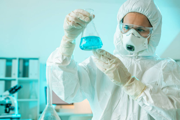 Young contemporary researcher in protective coveralls, respirator, eyeglasses and gloves studying blue liquid substance in tube - Foto, Bild