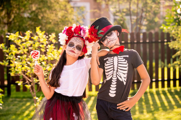 Dwoje uroczych dzieciaków z pomalowanymi twarzami stojących blisko siebie przed kamerą i patrzących na ciebie podczas świętowania Halloween - Zdjęcie, obraz