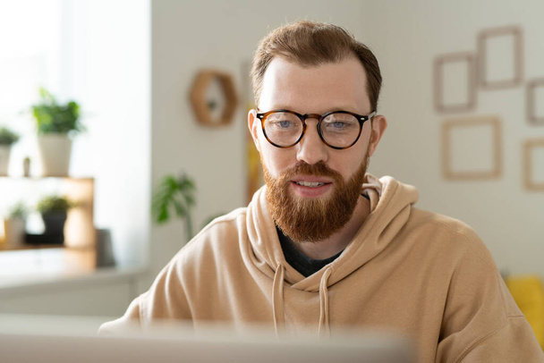 Junge bärtige freiberufliche Designerin mit Brille blickt auf Laptop-Display, während sie während der Fernarbeit durch Online-Informationen scrollt - Foto, Bild