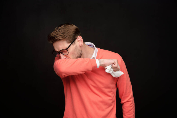 Zieke jongeman met bril tegen zwarte achtergrond en niezen in servet, virus concept - Foto, afbeelding