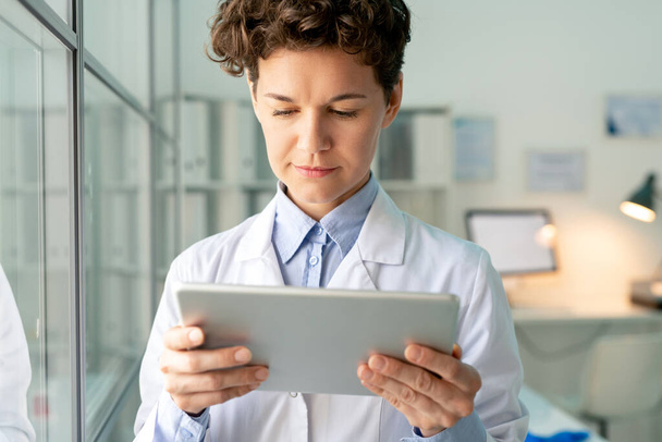 Junge ernstzunehmende Wissenschaftler oder Chemiker in weißem Kittel schauen sich Informationen auf dem Tablet-Bildschirm an, während sie im wissenschaftlichen Labor stehen - Foto, Bild