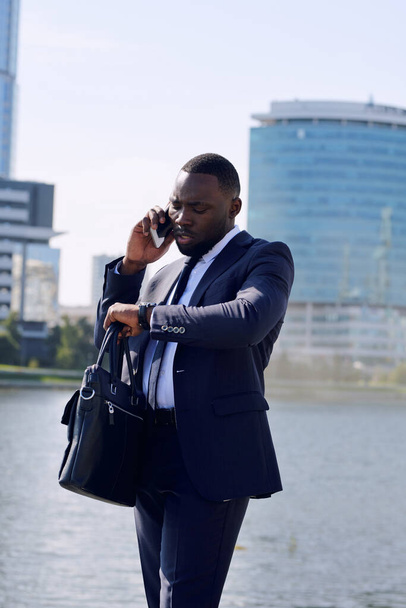 Jeune homme d'affaires africain élégant ou agent consultant client sur téléphone mobile au bord de la rivière tout en se tenant contre l'environnement urbain - Photo, image