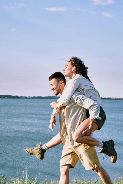Веселый молодой бородатый мужчина, дающий девушке кататься на спине на побережье, пока они веселятся во время похода - Фото, изображение