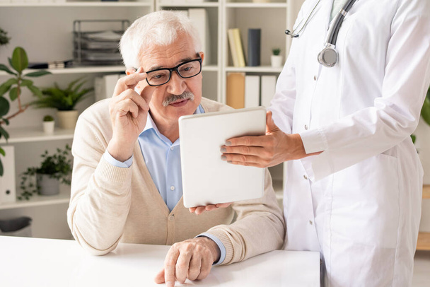 Szemüveges és alkalmi viseletű idős beteg, aki a fiatal orvos által a klinikákon fehérköpenyben tartott digitális tabletta kijelzőjén lévő egészségügyi adatokat nézi - Fotó, kép