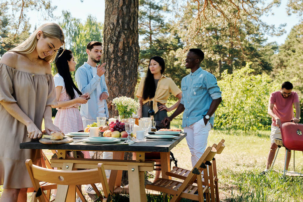 Grupo de jóvenes amigos internacionales en ropa de casualwear hablando por mesa servida bajo el pino mientras chica rubia cortando pan fresco para la cena - Foto, Imagen