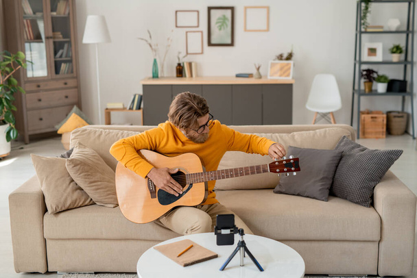 Joven músico barbudo o profesor de música en ropa casual sentado en el sofá en la sala de estar y tocando la guitarra delante de la cámara del teléfono inteligente - Foto, imagen